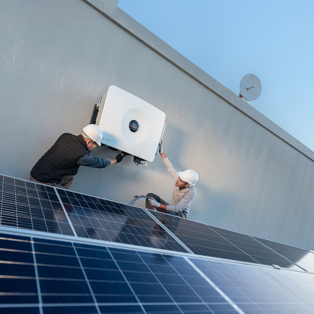Dwóch instalatorów montuje urządzenie Sunny Tripower 125 w ramach instalacji komercyjnej z modułami słonecznymi.