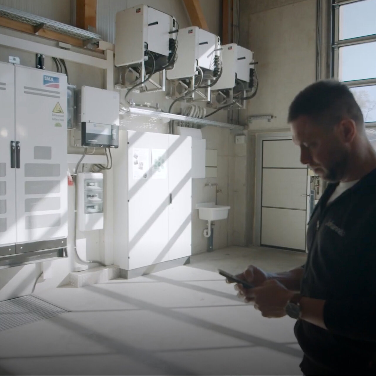 Duża hala, w której na ścianie wiszą liczne urządzenia SMA Sunny Tripower CORE1, szafa sterownicza SMA oraz dodatkowa szafa bezpiecznikowa; przed nimi stoi mężczyzna, który patrzy na swój telefon komórkowy.