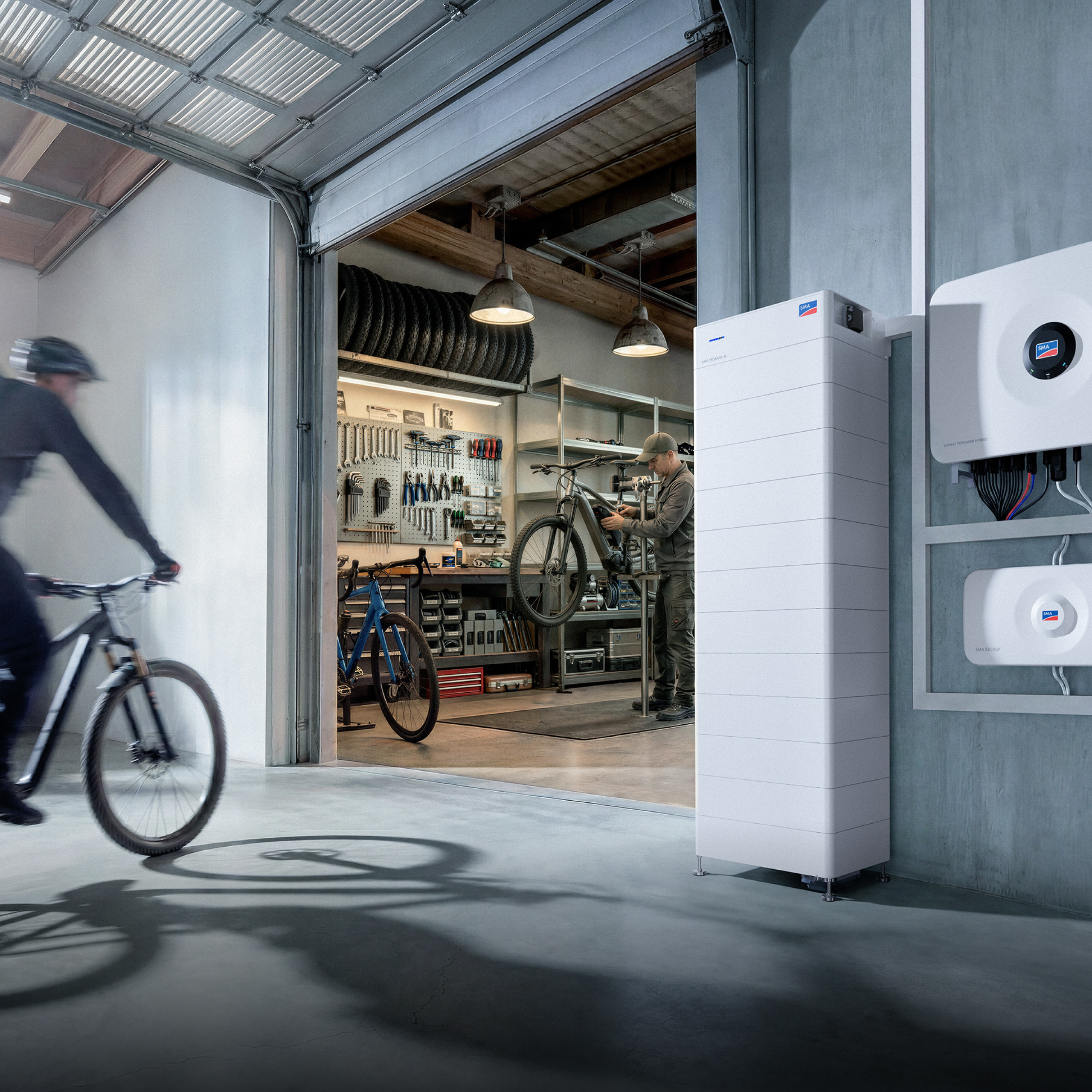 Warsztat rowerowy w hali magazynowej; przed nim stoi magazyn energii SMA Storage M, a na ścianie wiszą falownik Sunny Tripower Hybrid X oraz urządzenie SMA Backup.